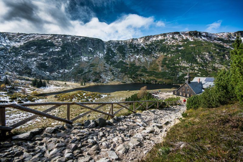 Zdjęcie wykonane ze szlaku pieszego przedstawia panoramę Karkonoszy, staw oraz drewniany budynek schroniska