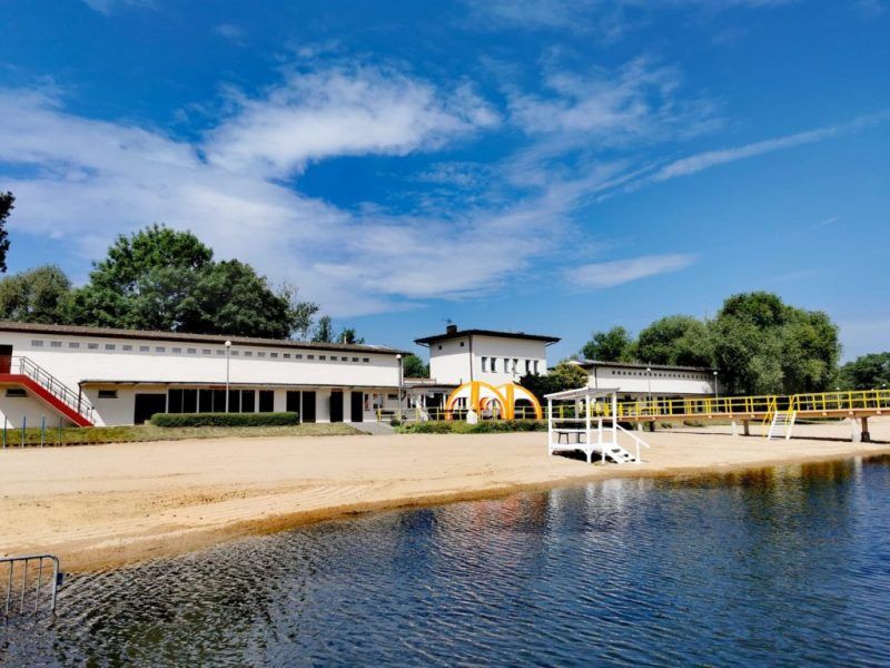 Na pierwszym planie zdjęcia widać kąpielisko łączące się z plażą. Na plaży znajduje się budka ratownika. Na drugim planie, w tle, znajdują się białe budynki kompleksu kąpieliska Kormoran