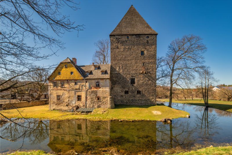 Solidna, prostokątna wieża z kamienia w stylu gotyckim, z wysokim, spadzistym dachem, otoczona fosą i przylegającym budynkiem mieszkalnym.