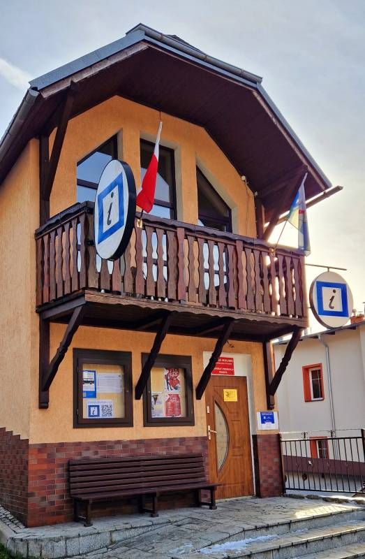 Wąski piętrowy budynek z nieproporcjonalnie dużym balkonem. Na budynku widać podwójne oznaczenie punktu informacji turystycznej oraz dwie flagi. Przy drzwiach wejściowych stoi ławka.