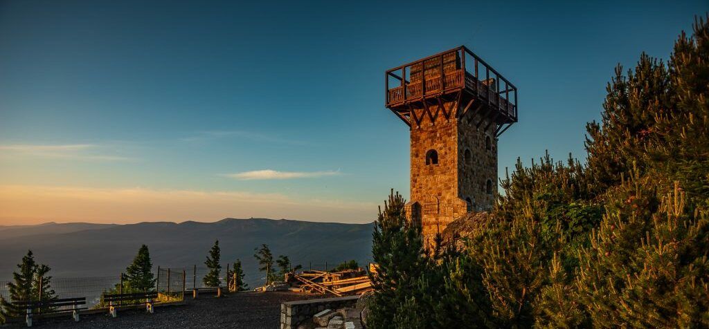 Kamienna wieża, z platformą widokową, wyłaniająca się zza krzewów. W tle panorama gór. Zachodża