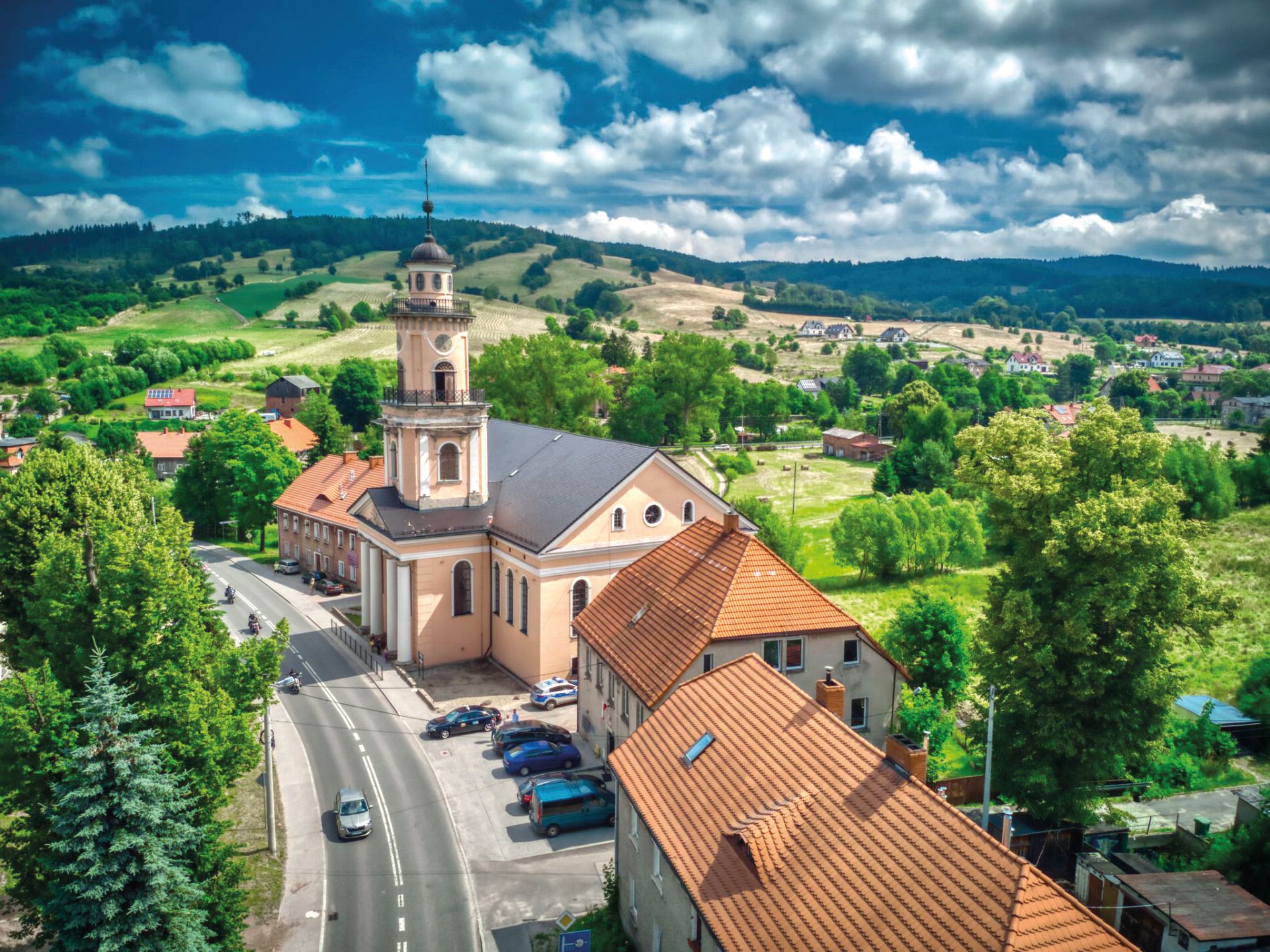 Widok na zabytkowy kościół przy ulicy. Widok na panoramę okolicy Głuszycy Górnej z pofałdowanymi, średniozalesionymi wzniesieniami w tle.
