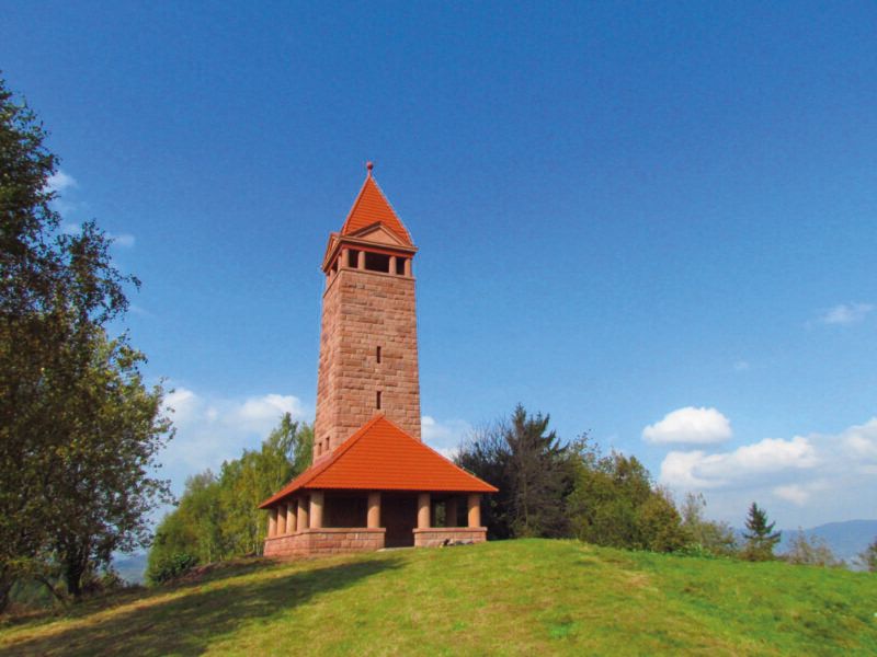 Widok na kamienną, zabytkową wieżę widokową o charakterystycznej czerwonej barwie na szczycie góry na tle błękitnego nieba.