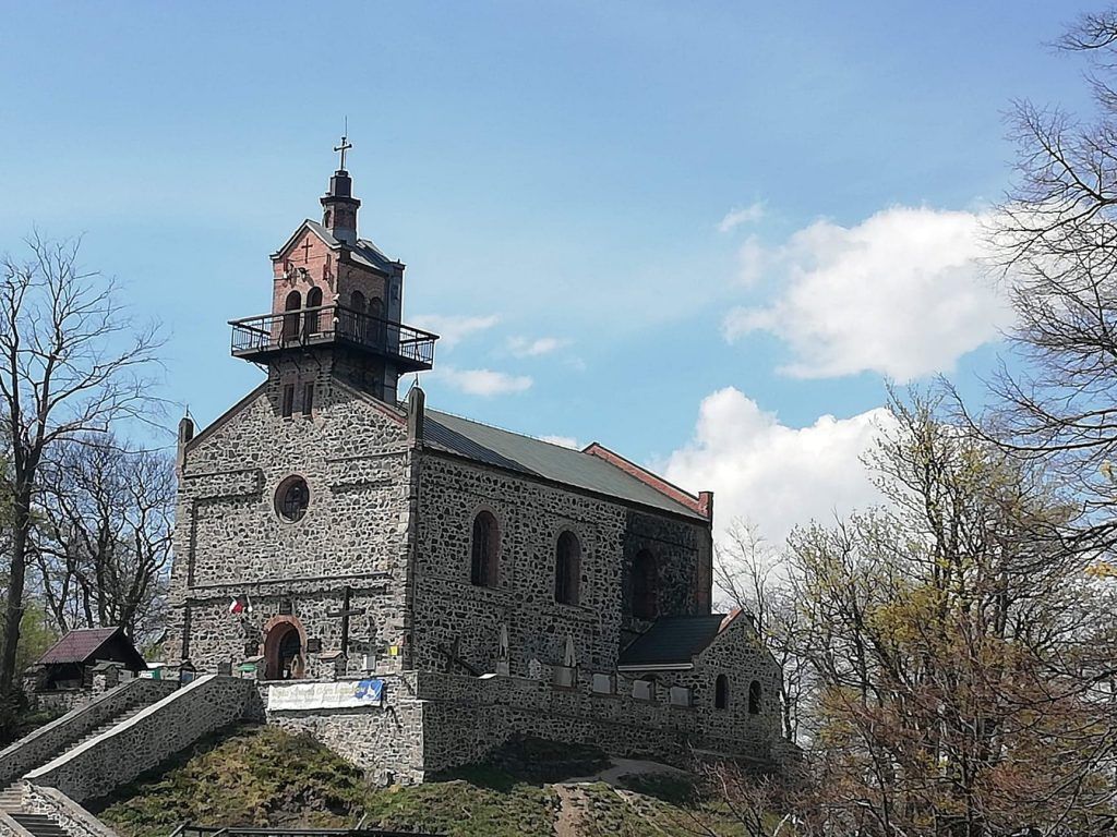 Widok na kamienny, nieotynkowany kościół z dwuspadowym dachem i niewielką wieżą w centralnej części fasady na tle nieba.