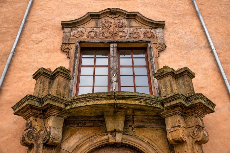 Na zdjęciu widać zbliżenie na pałacowe okno o ozdobnym, kamiennym obramowaniu – z delikatnymi zdobieniami i odbiciem drzew w szybie, które podkreśla historyczny charakter budowli.