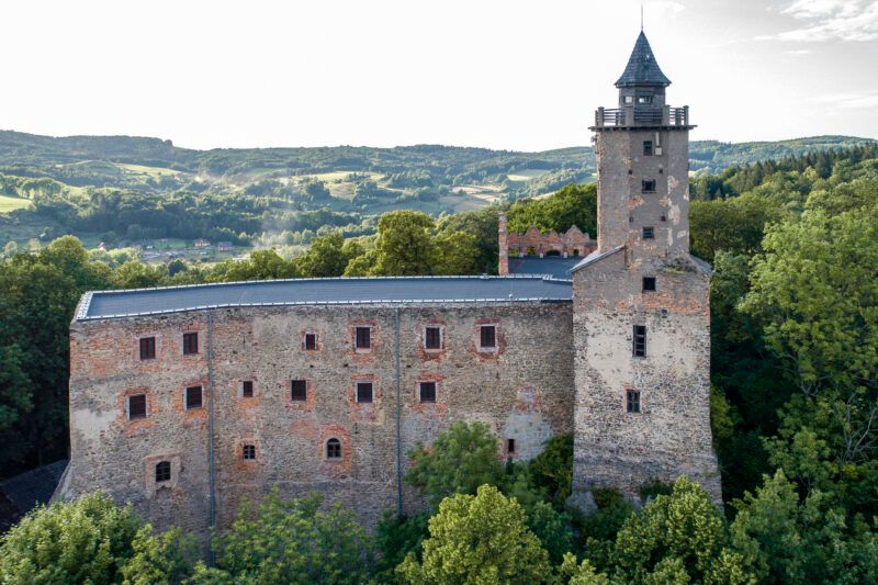 Zbliżenie na majestatyczne ruiny gotyckiego zamku, położone na wzniesieniu, w górzystym krajobrazie, wybijające się pośród zieleni drzew.