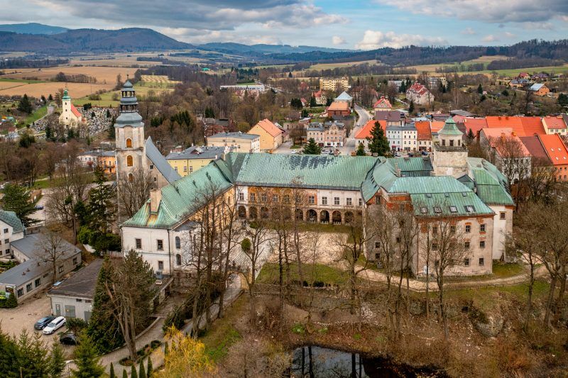 Widok z lotu ptaka na zamek Międzylesie ukazuje malowniczy kompleks historycznych budowli z czerwonymi dachami i wieżami, otoczony zielenią i wpisany harmonijnie w krajobraz górskiej doliny.