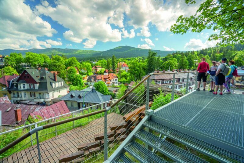 Widok na centrum Szklarskiej Poręby oraz Karkonosze w oddali przy wiosennej pogodzie z turystami widocznymi na platformie.