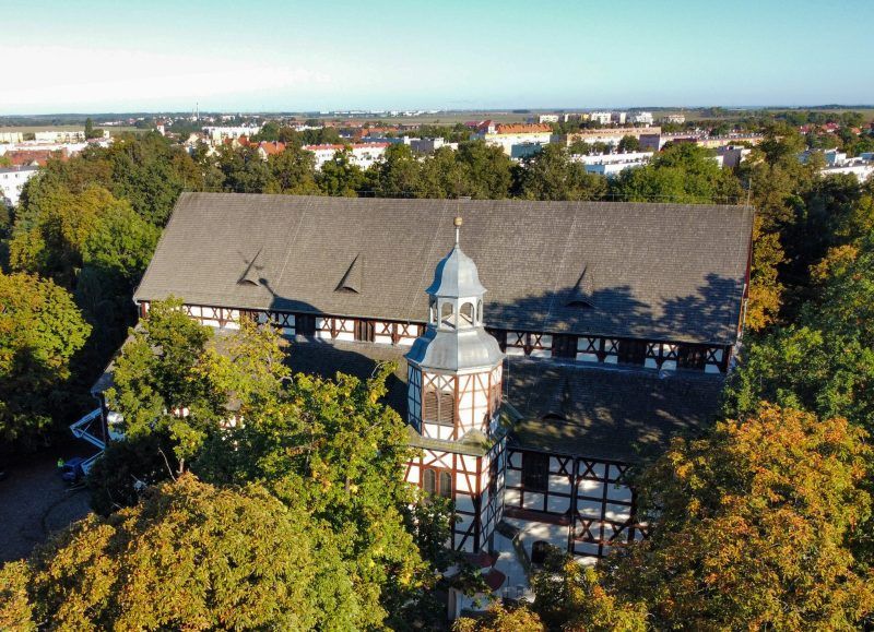 Zdjęcie z drona przedstawiające biało-brązową bryłę kościoła szachulcowego, wynurzającą się spośród drzew w parku,