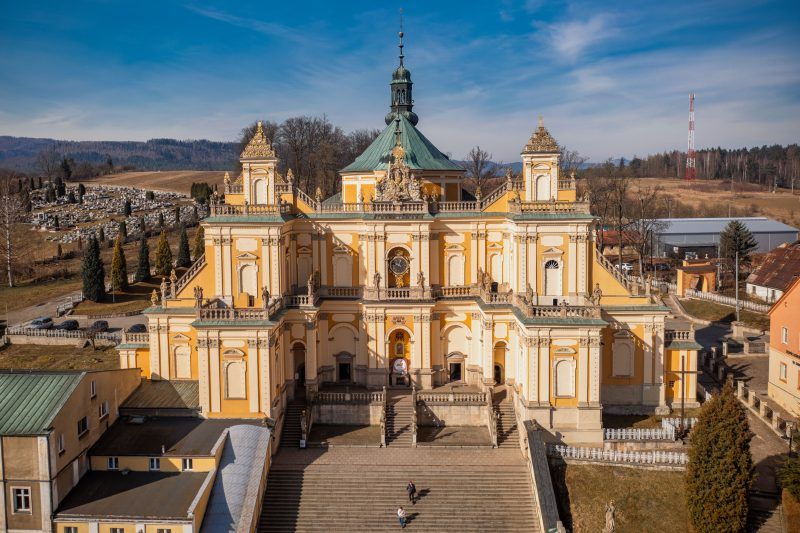 Na zdjęciu widać okazałe sanktuarium w Wambierzycach z monumentalnymi schodami prowadzącymi do barokowej fasady ozdobionej kolumnami i wieżami.