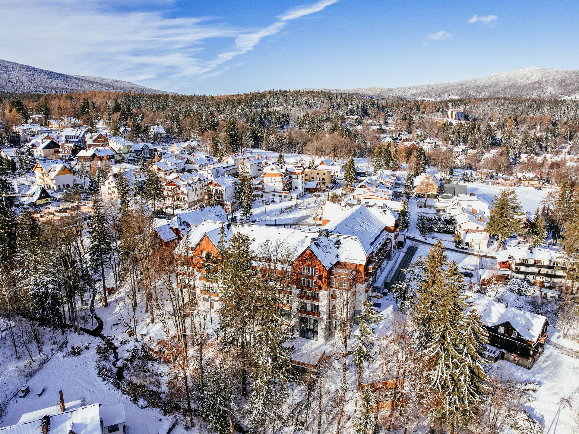 Zdjęcie z drona przedstawiające duży, wielopiętrowy budynek hotelu, na tle zimowego krajobrazu oraz panoramy gór.
