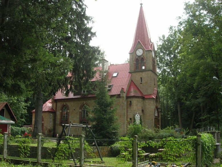 Schowany wśród gęstej roślinności budynek kościoła ze strzelistą wieżą z czerwonym dachem. Przed kościołem stoją ławki, huśtawka i piaskownica.
