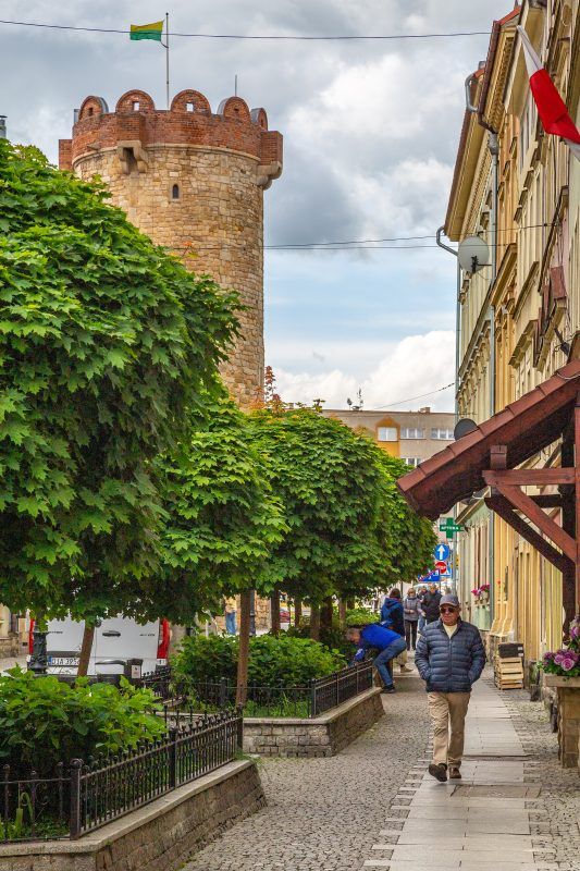 Zdjęcie ulicy w Złotoryi ze spacerującymi ludźmi oraz kamienną basztą z tle