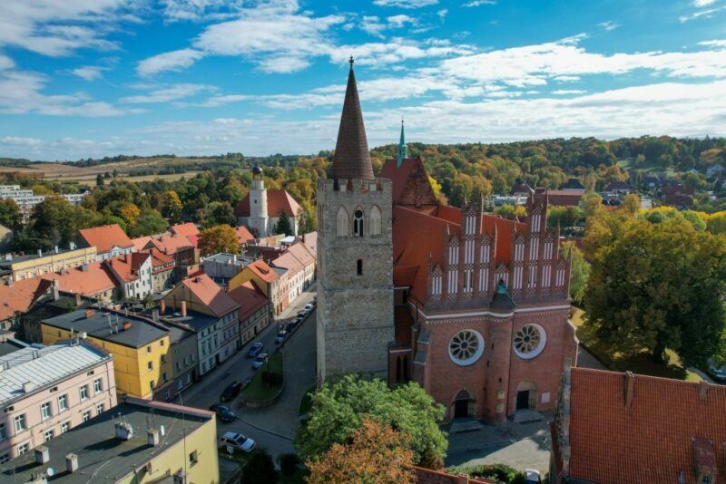 Widok z lotu ptaka na kościół, otoczony budynkami, w tle lasy oraz błękitne niebo.