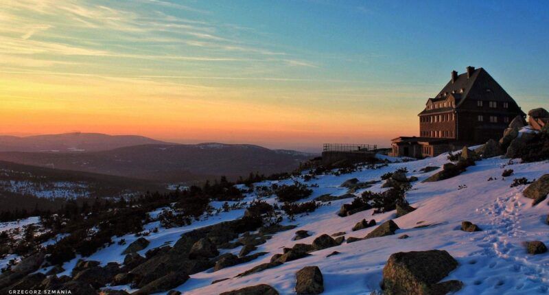 Górskie schronisko w zimowej szacie, położone na szczycie kamienistej góry, z piękną panoramą na sąsiednie wzniesienia.