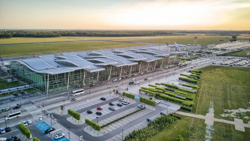 zdjęcie z lotu ptaka o zachodzie słońca, zdjęcie objemuje przeszklony port lotniczy wrocław, razem z częścią parkingu, oraz nawet kawałkiem pasu startowego samolotów