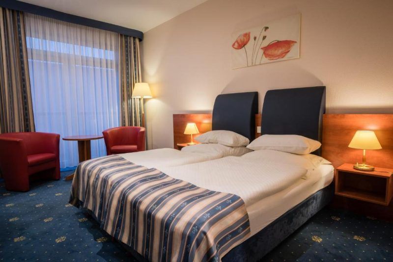 Pokój hotelowy z dużym łóżkiem podwójnym, szafkami nocnym i lampkami. Pod oknem okrągły stolik i dwa czerwone fotele. Na ścianie nad łóżkiem namalowane maki.