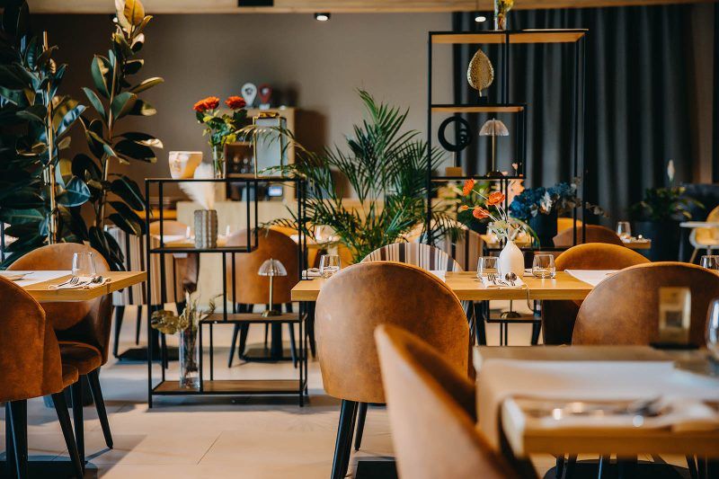 Nowoczesna, designerska restauracja hotelowa z elementami drewnianymi oraz metalowymi. Dekorację stanowią rośliny zielone, długie zasłony, drobne przedmioty ozdobne.