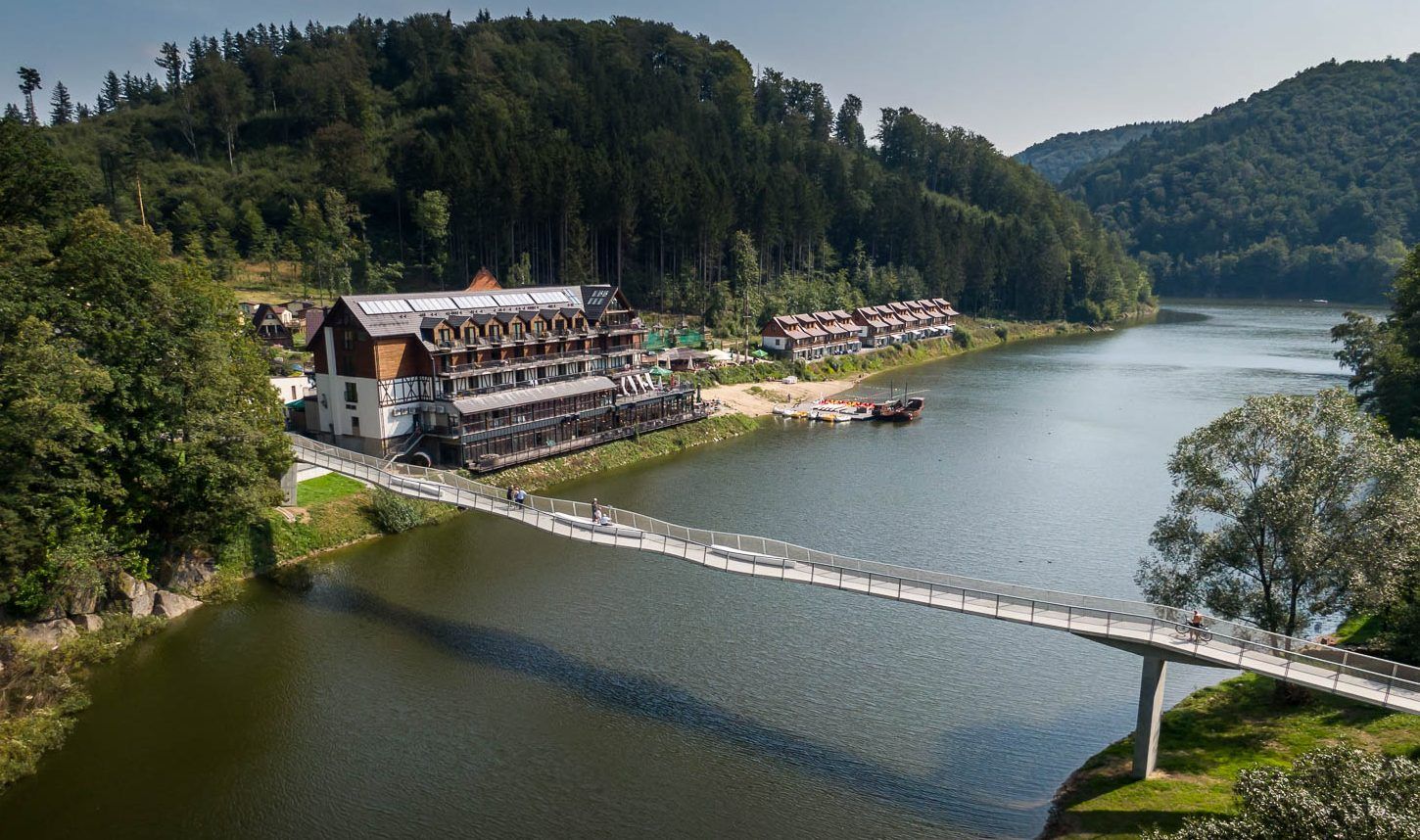 Kładka wstęgowa w Zagórzu Śląskim. Widok z drona na zbiornik wodny otoczony porośniętymi lasami wzgórzami. Na brzegu okazały obiekt hotelowy.