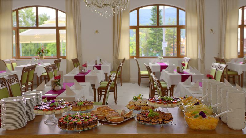 Przestronna, duża, jasna sala restauracyjna ze stołami nakrytymi białymi obrusami z bordowym bieżnikiem. Na pierwszym planie stół z deserami - ciasta, owoce w syropie, drobne wypieki.