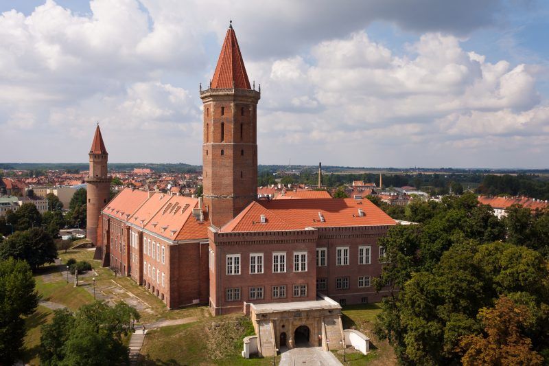 Panorama na budynek Zamku Piastowskiego z jedną wysoką i jedną niższa wieżą, dach pokryty czerwoną dachówką.