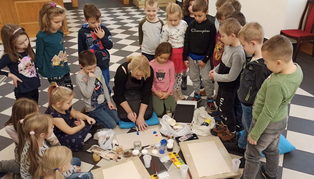 Kobieta pokazuje grupie dzieci jak malować farbkami