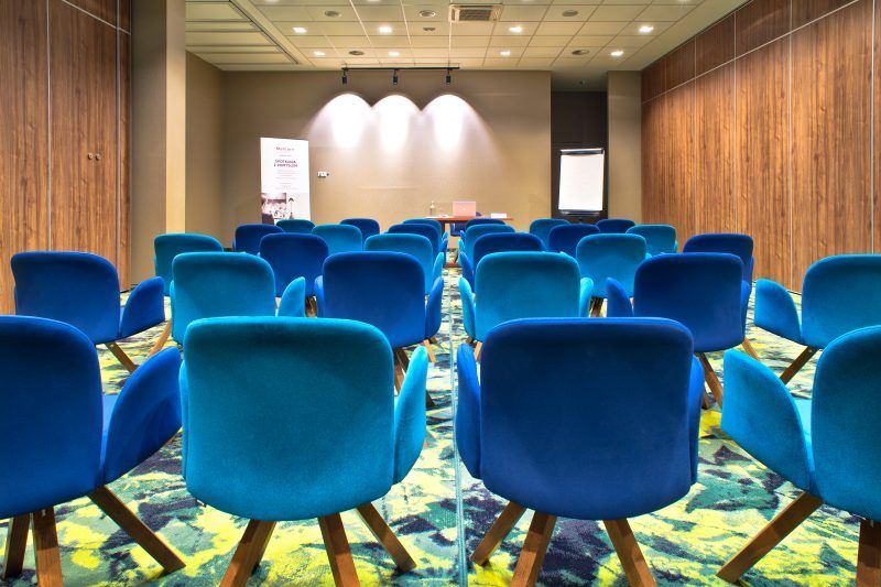 Sala konferencyjna. Na podłodze kolorowa jasna wykładzina. Ściany obite drewnem.