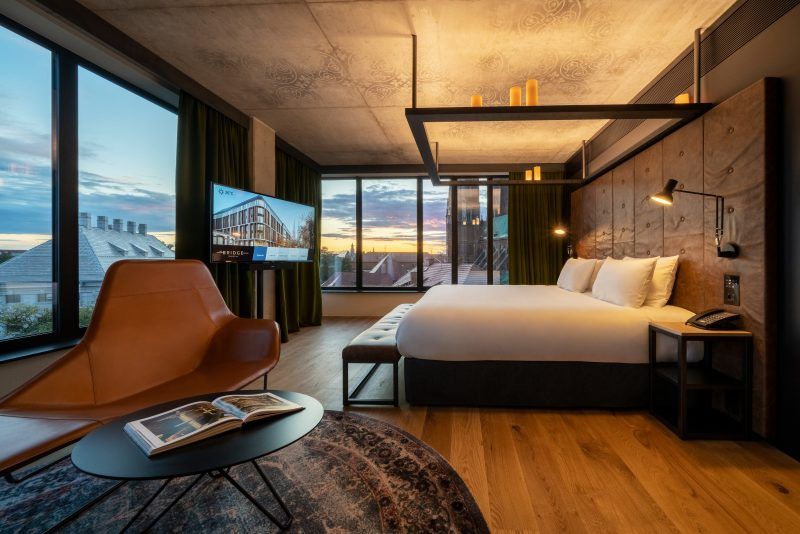 Zdjęcie przedstawia nowoczesny, elegancki pokój hotelowy urządzony w szarej, minimalistycznej stylistyce. Dominują w nim kolory – czerń, szarość i drewno. W pokoju znajdują się dwa łóżko typu king size z drewnianym zagłówkiem. Dwie ściany zostały przeszklone, roztacza się z nich piękny widok na zabytkowe budynki Ostrowa Tumskiego.