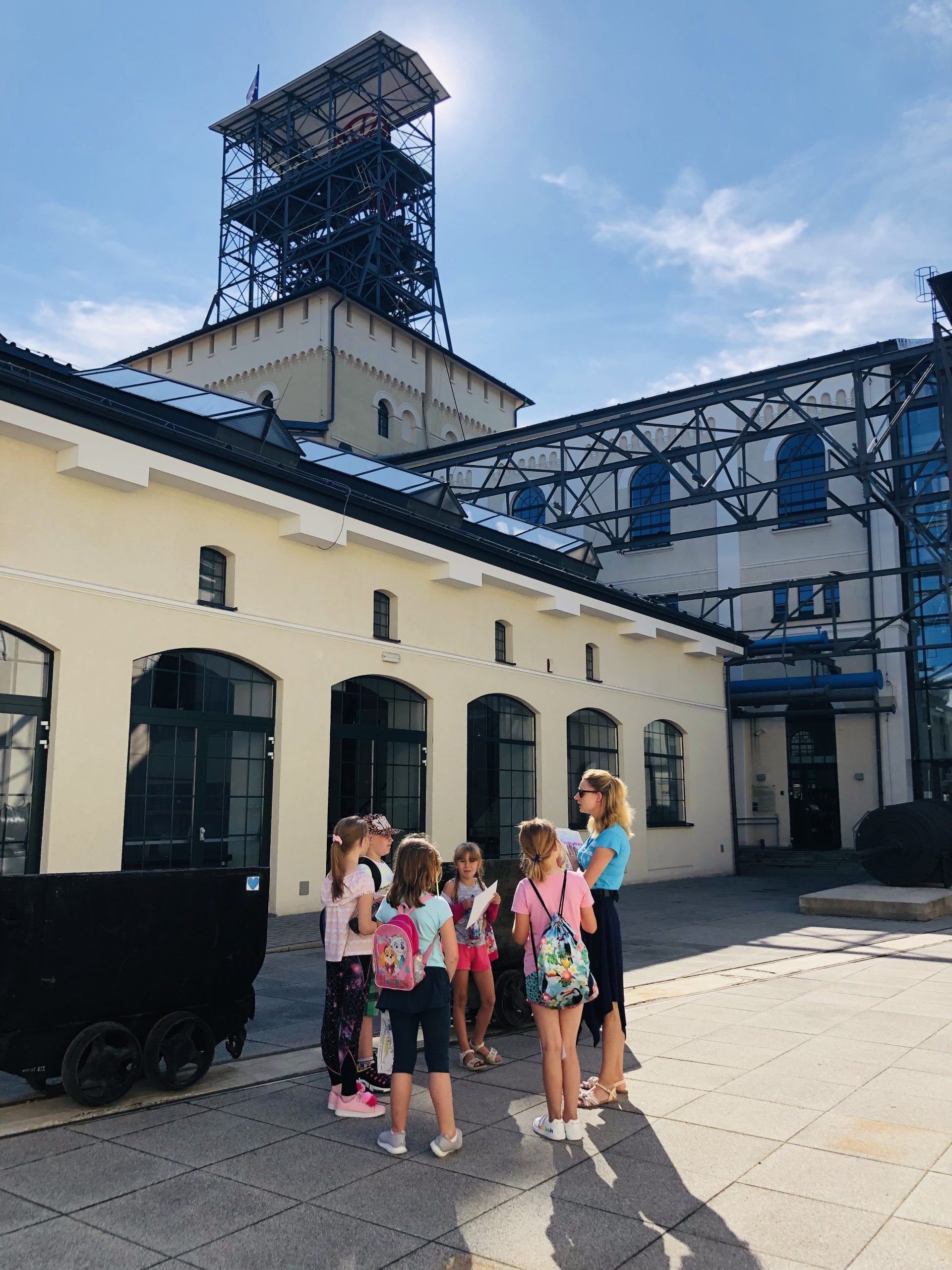 Grupka dzieci przez wejściem do zabytkowego budynku dawnej kopalni, w tle stoi szyb węglowy
