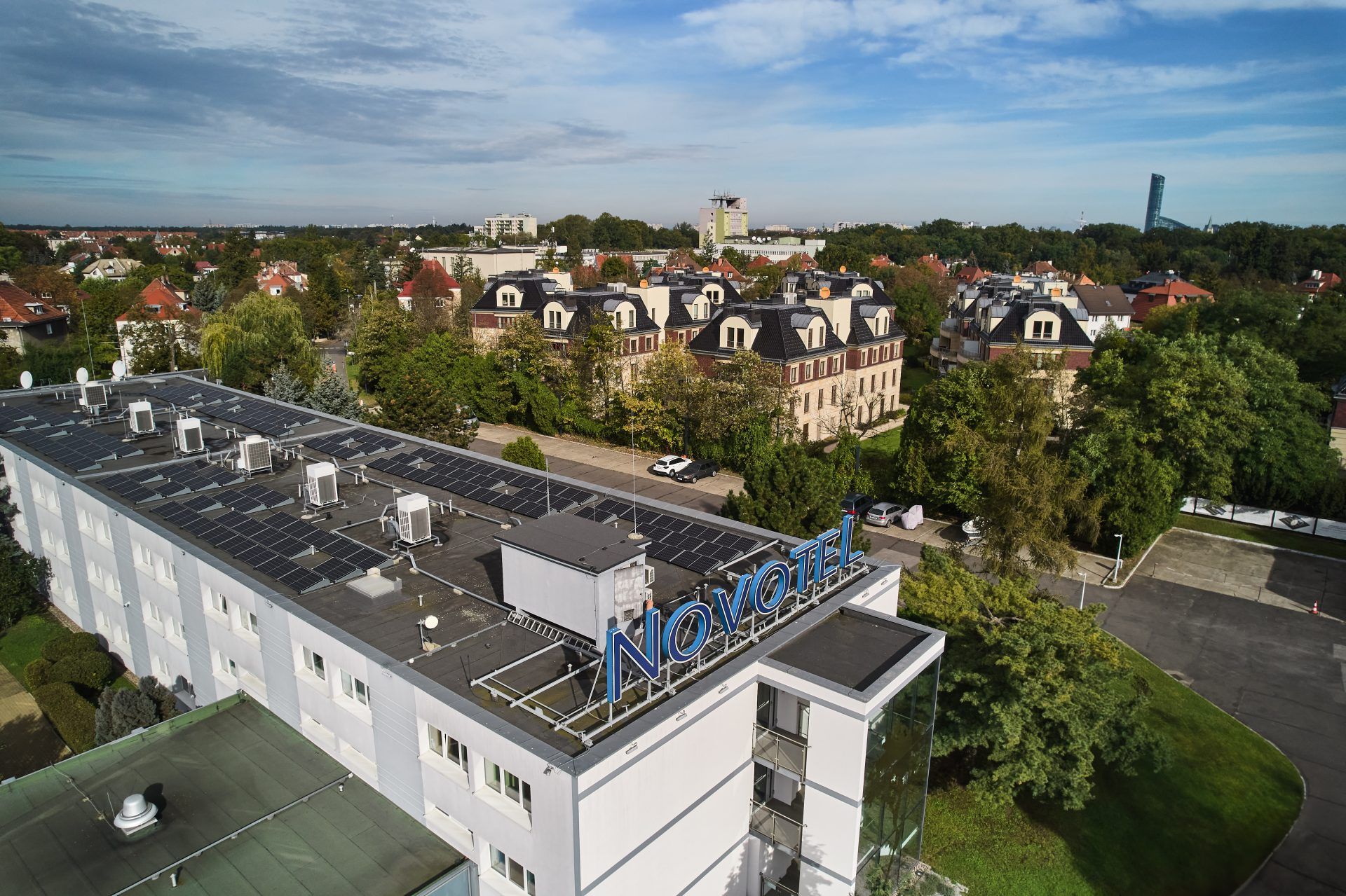 Widok z góry na budynek oznaczony niebieskim napisem „Novotel”, umieszczonym na dachu. W tle rozciągają się drzewa, osiedla i samotna wieża. Niebo jest lekko zachmurzone, nadając scenie spokojny nastrój. Hotel znajduje się pośrodku zielonego, miejskiego krajobrazu