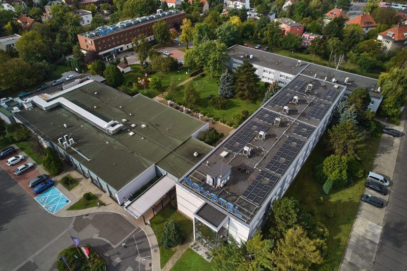 Zdjęcie ukazuje budynek hotelu otoczony zielenią. Przed hotelem znajdują się samochody oraz fragmenty trawnika. W tle widać zabudowania mieszkalne. Sceneria wygląda zadbanie i przyjaźnie.