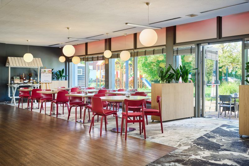 Zdjęcie pokazuje jasne pomieszczenie restauracyjne z czerwonymi krzesłami i rośliną w donicy ustawioną w rogu. Podłogę zdobi dywan, a przez okna widać zieleń na zewnątrz. Miejsce sprawia wrażenie przestronnego i spokojnego.