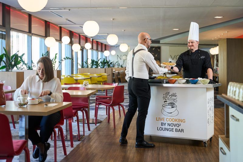Na fotografii kelner i kucharz podają jedzenie klientowi siedzącemu przy stoliku w restauracji. W tle widoczne są dekoracje wnętrza i kolejne stoliki. Scena ukazuje moment obsługi gościa w przyjaznej, żywej atmosferze.