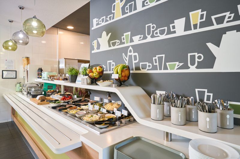 Obraz ukazuje hotelowy bufet śniadaniowy, pełen różnorodnych potraw. Na ladzie znajdują się misy z płatkami, pojemniki z napojami, pieczywo i dodatki śniadaniowe. Nad bufetem zawieszono ozdobne lampy, a na ścianie dominuje duża grafika z filiżankami, dzbankami i kubkami w białym kolorze na ciemnym tle. Całość robi wrażenie czystości, porządku i gościnności.