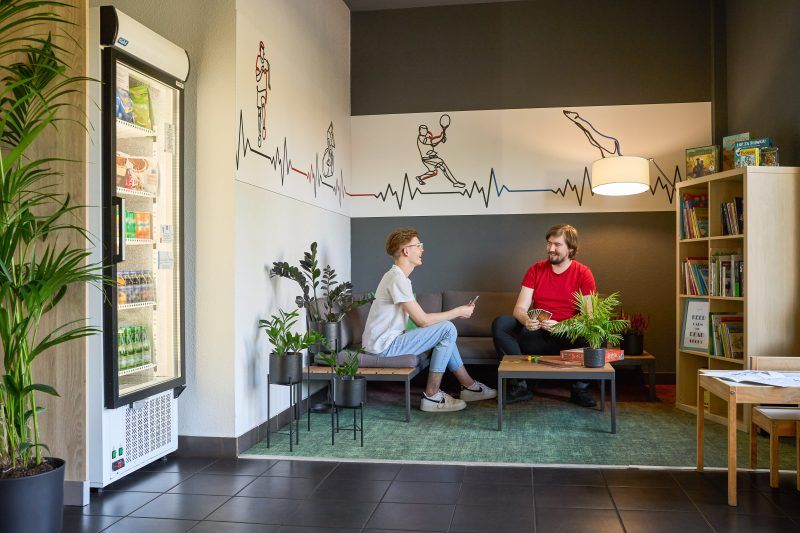 Zdjęcie pokazuje niewielki, przytulny kącik wypoczynkowy wewnątrz hotelu. Dwie osoby siedzą naprzeciwko siebie na wygodnych fotelach i rozmawiają. Ściana za nimi jest ozdobiona dużą grafiką w stylu line-art, przedstawiającą zarys gór i ludzi wspinających się na szczyt. Obok foteli stoją rośliny i drewniany stolik, a całość wnętrza zachęca do relaksu.
