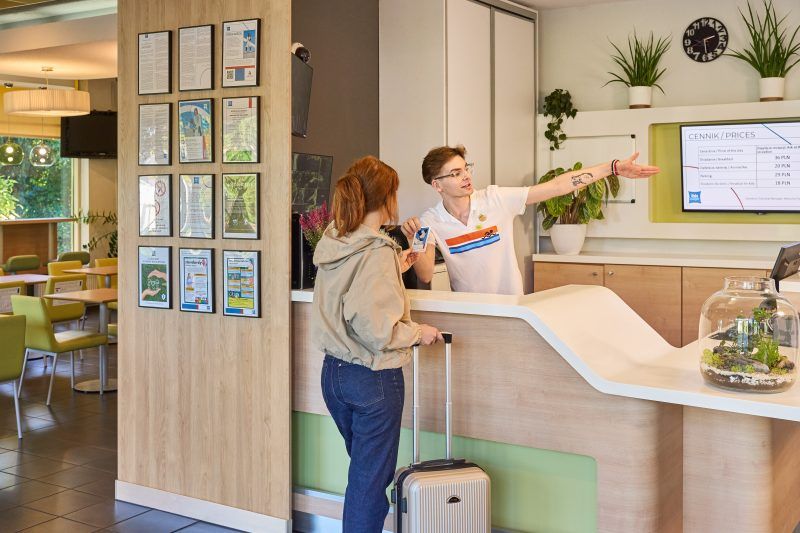 Fotografia przedstawia przestrzeń recepcji hotelowej, urządzoną w jasnych, przyjaznych kolorach. Przy ladzie recepcyjnej stoi recepcjonista w białej koszuli, rozmawiający z kobietą w dżinsach i beżowej bluzie, która trzyma walizkę. W tle na ścianie wisi ekran z informacjami, a wokół recepcji znajdują się rośliny doniczkowe i drewniane wykończenia wnętrza, co tworzy nowoczesną i ciepłą atmosferę.