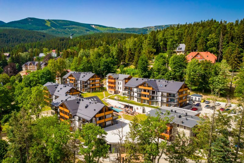 kompleks apartamentowy położony wśród zieleni u podnóża gór, otoczony lasem i górami w tle. Budynki mają drewniane akcenty elewacyjne, a w centralnej części znajduje się basen z leżakami.