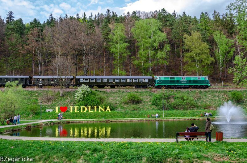 Zdjęcie przedstawia pociąg jadący po nasypie graniczącym ze stawem i widocznym napisem "I love Jedlina"