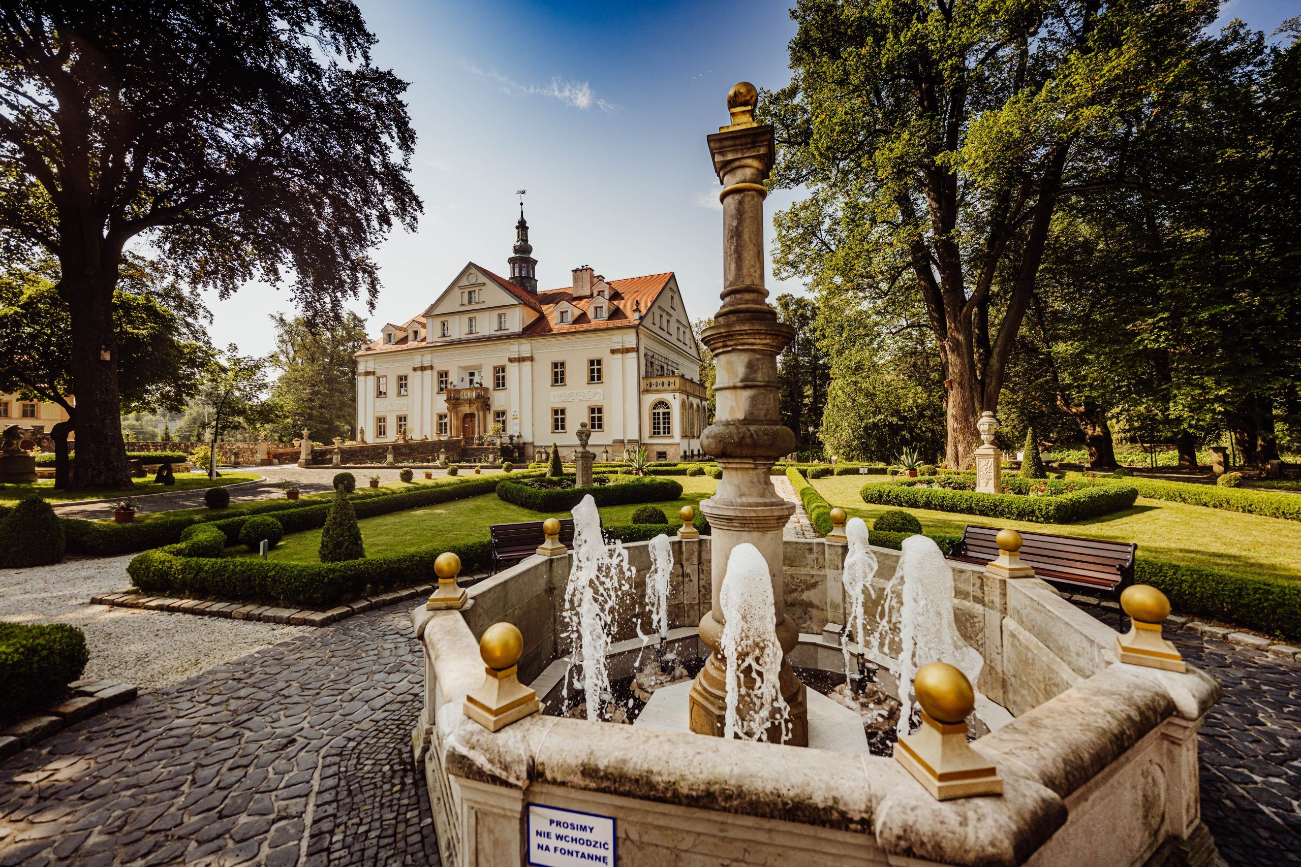 Zdjęcie przedstawiające Pałac Ciechanowice, ogród oraz czynna fontannę, którą widać na pierwszym planie. Zdjęcie wykonane przy słonecznej pogodzie.
