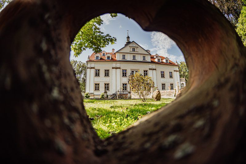 Zdjęcie wykonane zza otworu w murze, który to otwór jest w kształcie serca. W kadrze znajduje się fasada Pałacu Ciechanowice.
