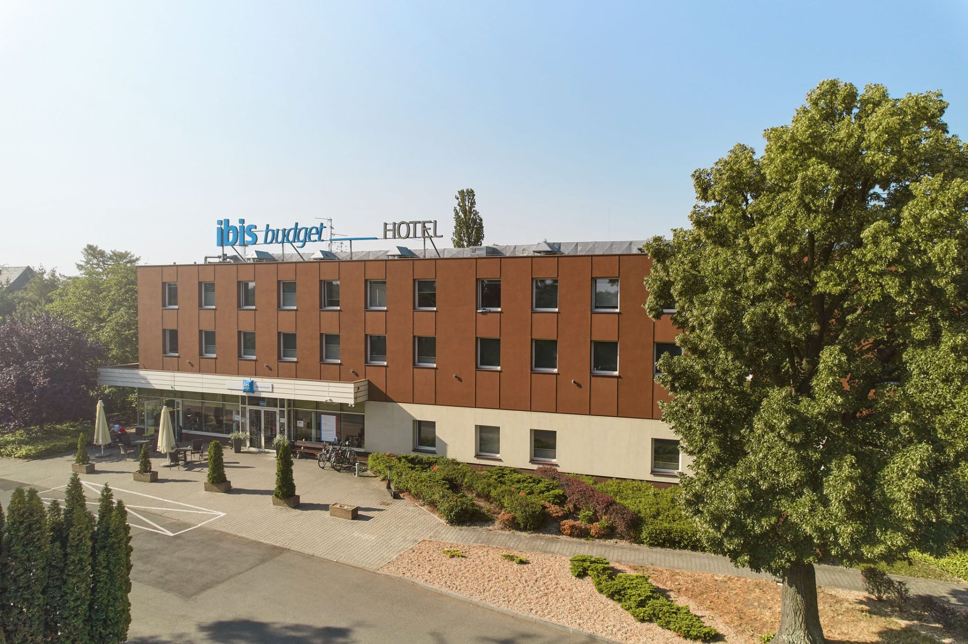 Na zdjęciu widać zewnętrzną fasadę hotelu ibis budget, zbudowaną z czerwonej cegły. Budynek ma kilka pięter, a jego bryła jest prosta i nowoczesna. Przed wejściem znajdują się starannie przycięte krzewy oraz podjazd, na którym zaparkowano kilka samochodów. W tle rosną wysokie drzewa, co nadaje miejscu spokojny, zielony charakter.