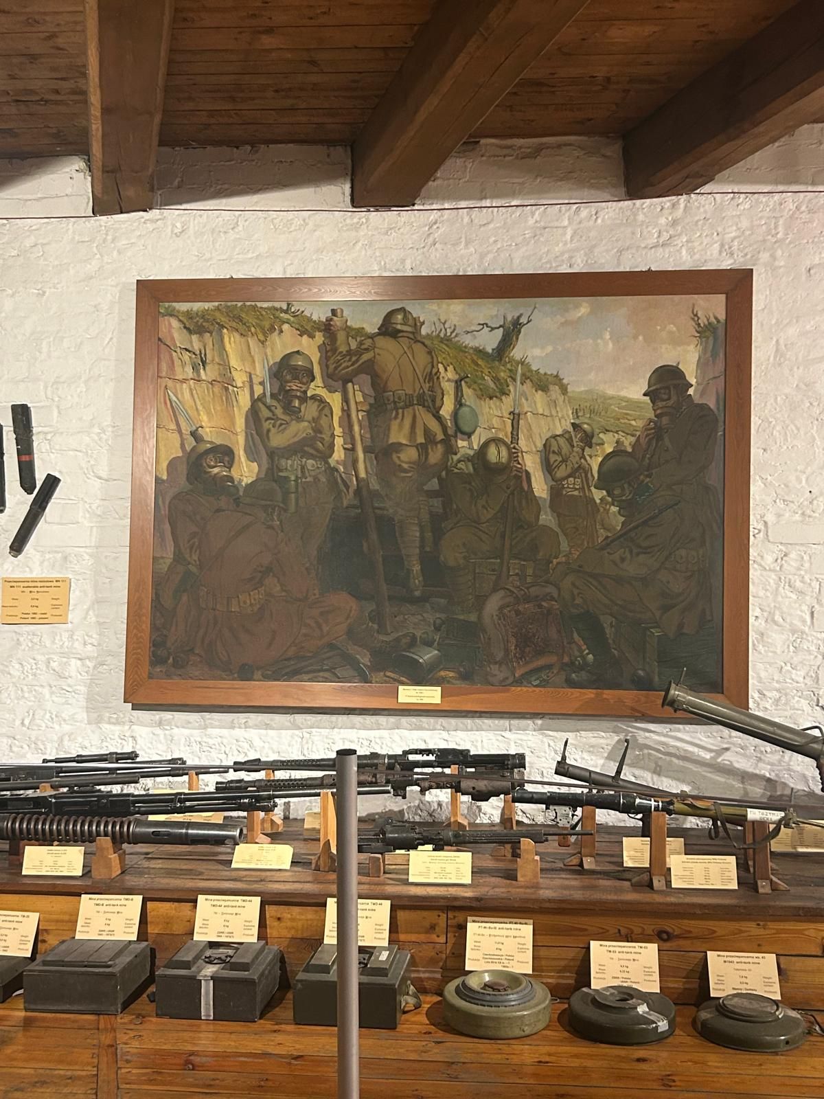 Zdjęcie przedstawia obraz żołnierzy znajdujących się w okopie. Pod obrazem ustawiona jest broń palna oraz miny