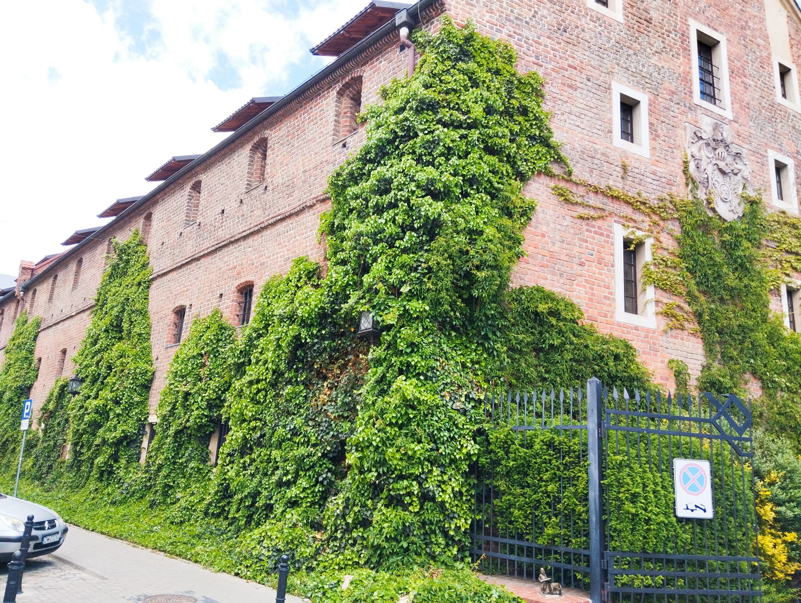 Zdjęcie przedstawia budynek z oknami wykonany z czerwonej cegły obrośnięty rośillnością. Na pierwszym planie widać bramę oraz samochód.