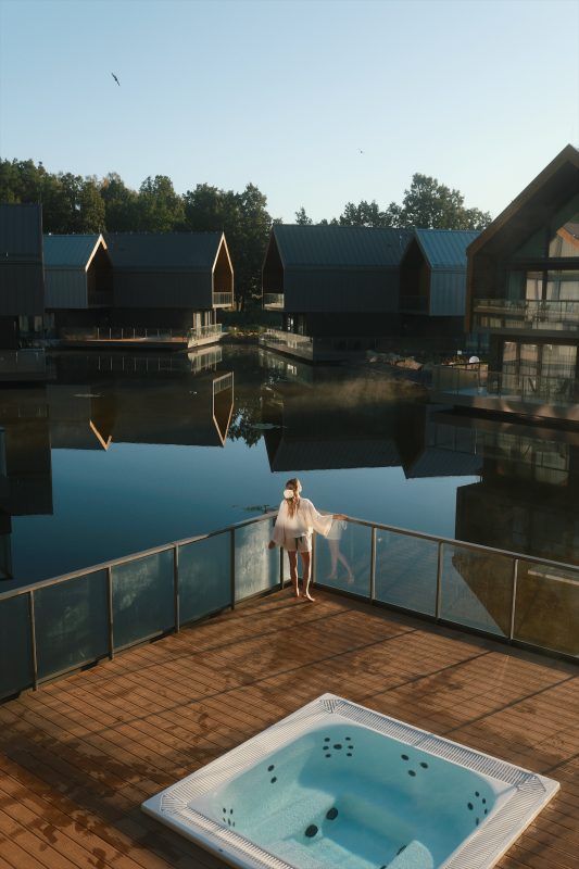 pionowe zdjęcie z tarasu pokoju hotelowego na którym znajduję się jacuzzi. Na tarasie stoi koboieta patrząca na wodę w której odbijają się inne pokoje hotelowe
