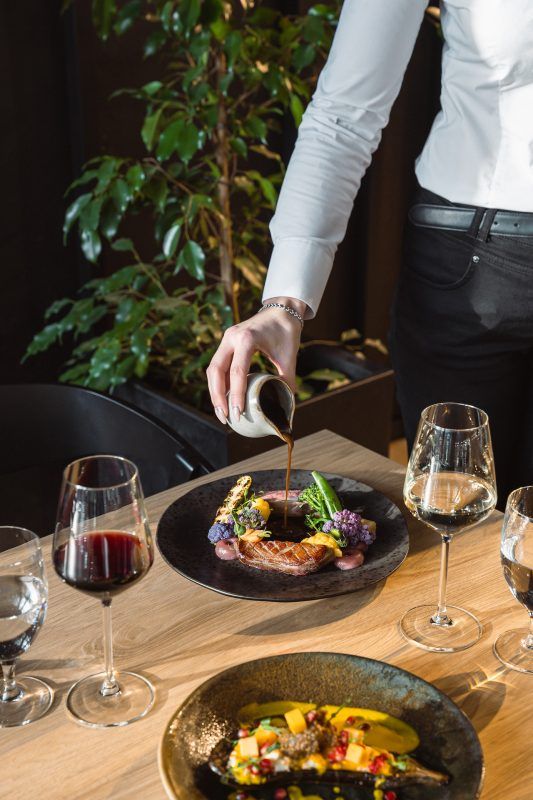 Na zdjęciu widać elegancko nakryty stół z daniami kuchni fine dining oraz osobę polewającą sosem danie główne. Na stole znajdują się również kieliszki z czerwonym i białym winem oraz szklanki z wodą.