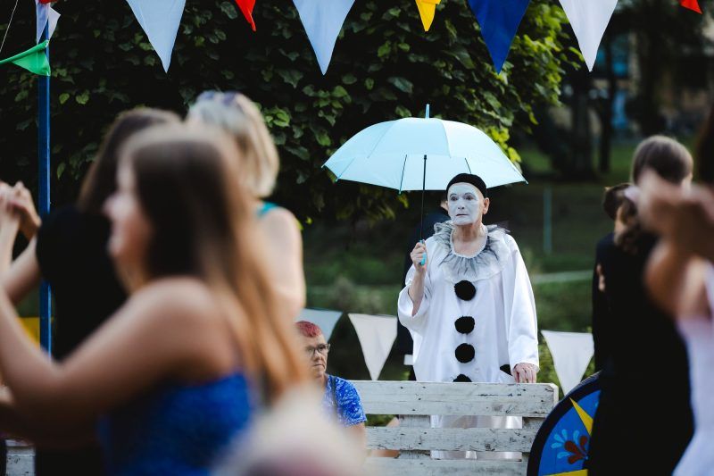 Na pierwszym planie stoi kobieta w niebiejskiej koszulce na ramiączkach odwrócona do obiektywu plecami a obok niej wyłania się obraz kobiety ubranej na biało z białym parasolem w ręku.