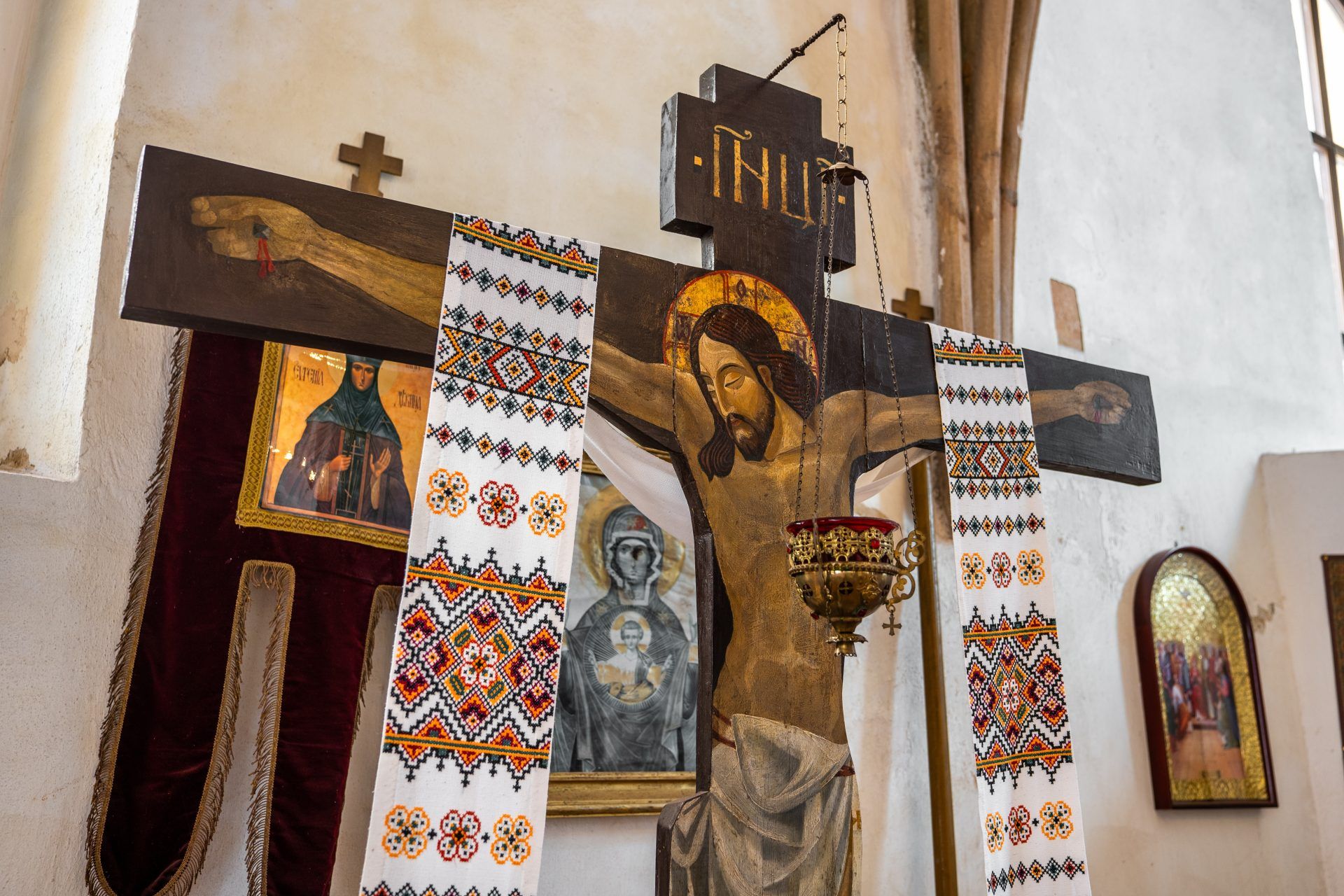 Drewniany krucyfiks w stylu bizantyjskim, z realistycznie przedstawioną postacią Chrystusa – smukłą, z wyraźnie zaznaczonymi ranami, głową pochyloną w lewo i złotą aureolą wokół głowy, zawieszony na tle ozdobnego ikonostasu.