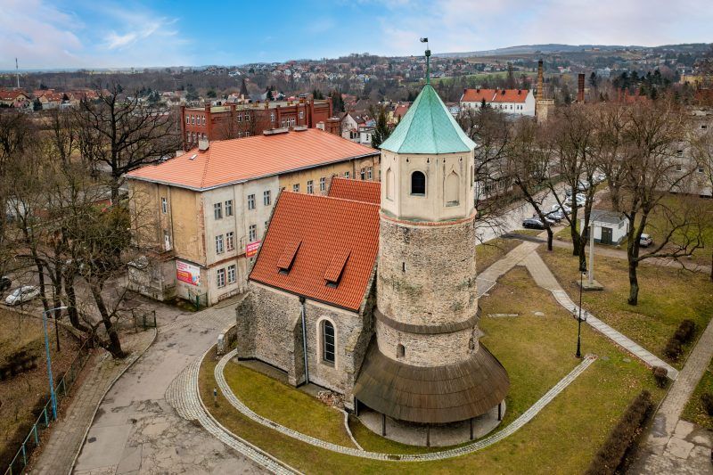 Widok z lotu ptaka na niewielką, romańską budowlę z kamienia polnego, o prostych, surowych formach, z charakterystyczną półkolistą apsydą i masywną bryłą na planie prostokąta.