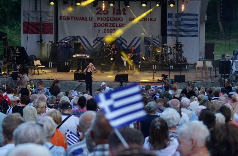 Zdjęcie sceny z instrumentami i publicznością. Na pierwszym planie widoczna jest grecka flaga.