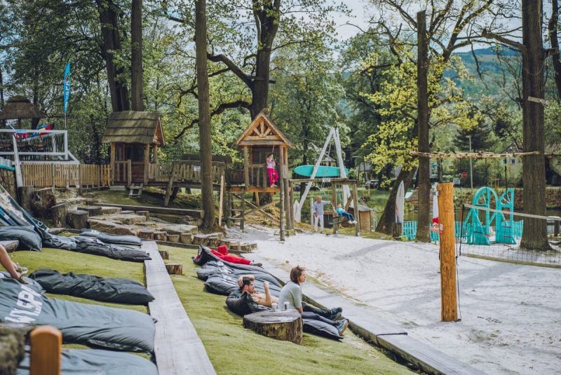 Zdjęcie przedstawiające plażę i plac zabaw z drewnianymi domkami i turystami wypoczywającymi na skraju zbiornika